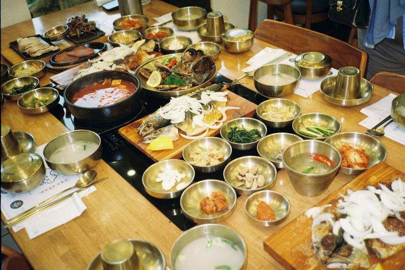 나무 테이블 위에 놋그릇 반찬 여러 가지와 찌개, 생선구이가 차려진 상