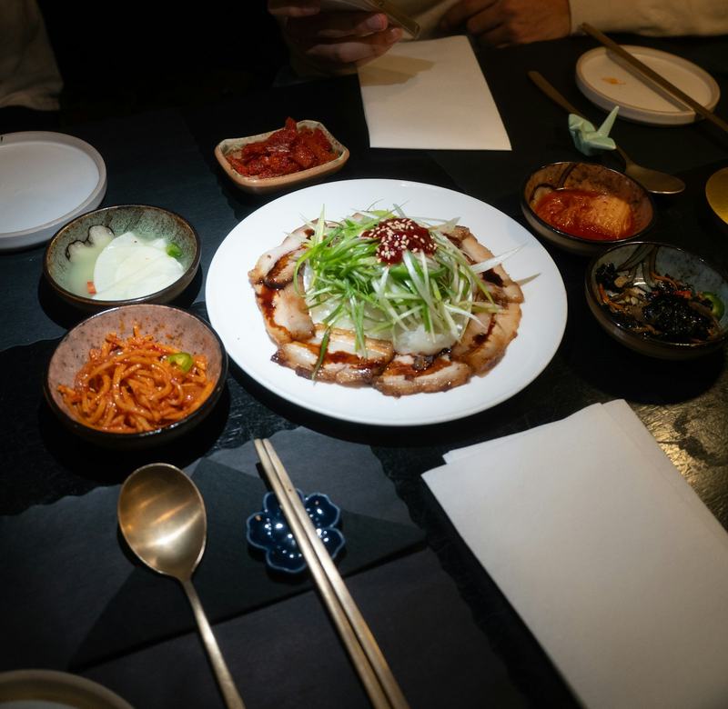 어두운 테이블 위 흰 접시에 담긴 보쌈과 대파채, 주변에 반찬 그릇
